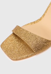 Primer plano de una sandalia dorada brillante con punta abierta cuadrada y una sola correa en la parte frontal.