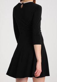 Abito nero con maniche lunghe, gonna svasata e dettaglio a oblò sul retro. Realizzato in tessuto morbido con una rifinitura bianca al collo, dotato di una vita aderente.