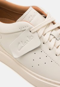 White leather sneaker with perforated details on the toe, matching laces, and a textured tongue featuring "Clarks" branding.