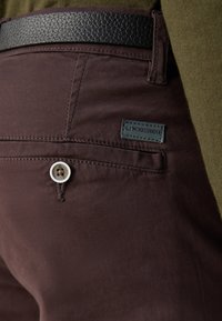 Καφέ παντελόνι από ύφασμα διαθέτει μια μαύρη δερμάτινη ζώνη, κλείσιμο με κουμπί μπροστά και μια μικρή επωνυμία κοντά στην πίσω τσέπη.