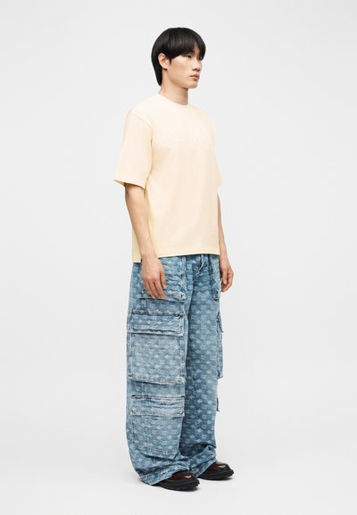 Νεαρός άνδρας που στέκεται πλαγιαστά φορώντας μια ανοιχτό μπεζ, υπερμεγέθη μπλούζα και φαρδιά μπλε παντελόνια cargo με μεγάλες τσέπες και μαύρα παπούτσια.