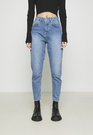 Woman wearing high-waist light blue jeans, black cropped ribbed long sleeve top, and black ankle boots, standing against a plain background.