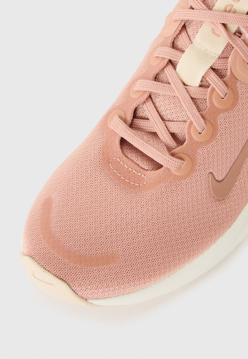 Zapatilla deportiva de color rosa claro con parte superior de malla, detalles texturizados y suela acolchada blanca. Presenta puntera redonda y diseño de cordones.
