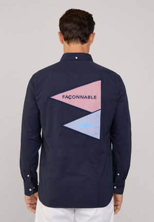 Camicia a maniche lunghe blu navy con un design triangolare sul retro realizzato in tessuto a strisce rosse e blu, con testo in rilievo "Faconnable" e dettaglio del pesce.