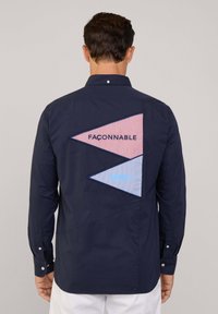 Marine blouson met een driehoekig ontwerp op de achterkant, gemaakt van rood en blauw gestreepte stof, met gegraveerde tekst "Faconnable" en een visdetail.