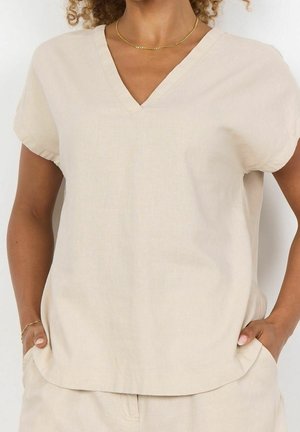 Vrouw draagt een beige V-hals T-shirt met korte mouwen en bijpassende broek, met handen in de zakken, zichtbare gouden ketting en armband.