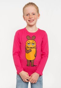 Lächelndes Mädchen mit roten Haaren und Sommersprossen, das ein leuchtend pinkes Langarmshirt mit einer Cartoon-Maus-Illustration und eine blaue Jeans trägt.