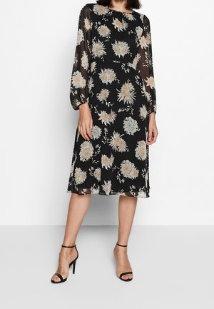Vrouw draagt een zwarte midi-jurk met beige en witte bloemenprints, doorzichtige lange mouwen en zwarte sandaaltjes met hoge hak en enkelbandje.