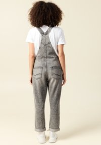 Pantalones de mezclilla gris con tirantes ajustables, puños enrollados y dos bolsillos traseros, mostrados desde la parte de atrás contra un fondo neutro.