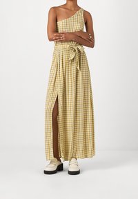 Vrouw gekleed in een geel-wit geruit één-schouderjurk met een tailleband en een zijsplit, in combinatie met crèmekleurige instappers.