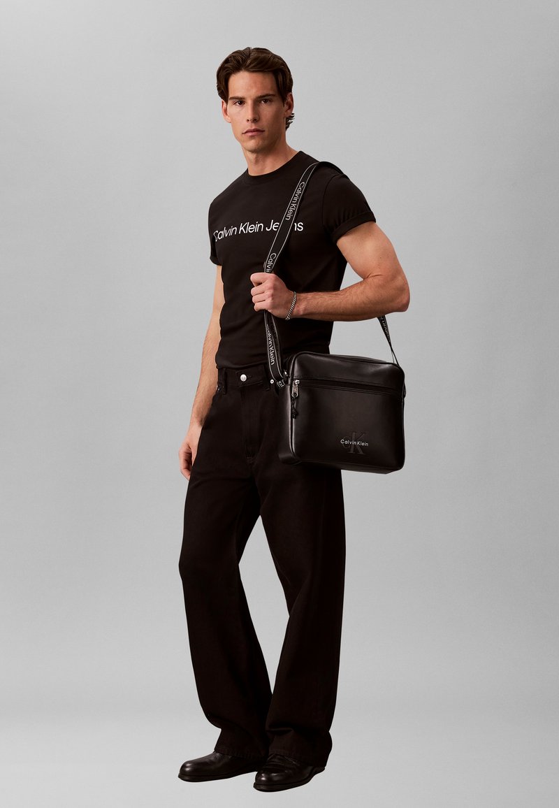 Άνδρας με μαύρο T-shirt Calvin Klein Jeans και μαύρο παντελόνι, κρατώντας μία μαύρη τσάντα ώμου Calvin Klein με λουράκι λογότυπο.
