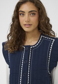 Gilet trapuntato blu navy con scollo rotondo, tre grandi bottoni e cuciture a zigzag bianche a contrasto lungo i bordi. Tessuto strutturato.