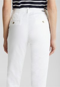 Pantalon en coton blanc à coupe slim, avec des poches arrière ornées de boutons dorés et une ceinture épurée. Sans motifs ni textures apparents.