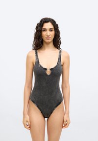 Γυναίκα  με σγουρά μαλλιά που φοράει αμάνικο σκούρο γκρι κορμάκι με ριγέ υφή, με άνοιγμα στο μπροστινό μέρος και κούμπωμα, σε απλό ανοιχτό φόντο.