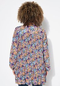 Květinové šaty s dlouhým rukávem, které mají skládací záda, zářivé barvy včetně fialové, oranžové a modré, s texturovaným vzorem na látce.