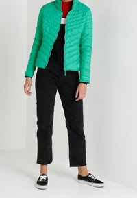 Zielona pikowana kurtka puchowa z wysokim kołnierzem, noszona na czarnym swetrze w czerwone paski i czarnych skróconych dżinsach, zestawiona z czarnymi trampkami.