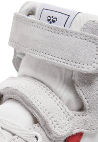 Szary zamszowy tenisówka z białą podszewką z tkaniny, posiada rzep, przeszycia jako akcenty oraz czerwony detal w pobliżu podeszwy.