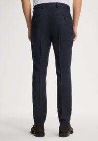 Nachhaltig dunkelblaue maßgeschneiderte Hose mit kariertem Muster, glatter Textur und zwei Gesäßtaschen mit Knopfverschlüssen. Kombiniert mit braunen Knöchelschaftstiefeln.
