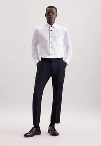 Wit langärmelig overhemd met knopen en een zak, gecombineerd met donker gesneden broek en zwarte schoenen, tegen een neutrale achtergrond.