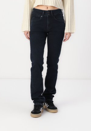 Dark blue, slim-fit jeans with a mid-rise waist and two-button closure; worn with black shoes featuring white stripes and a gum sole.
