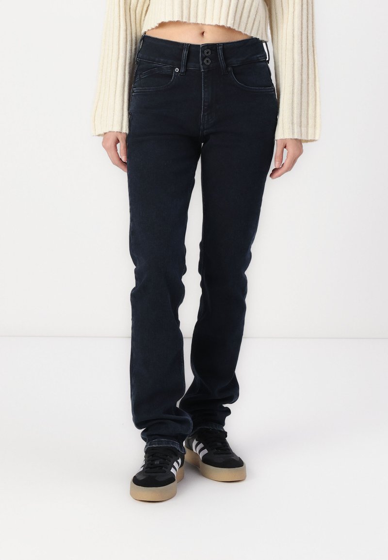 Dark blue, slim-fit jeans with a mid-rise waist and two-button closure; worn with black shoes featuring white stripes and a gum sole.