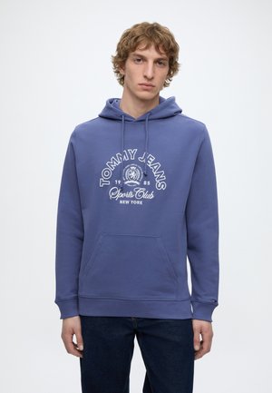 Jonge man draagt een blauwe Tommy Jeans Sports Club hoodie met een voorzak en trekkoorden, staand tegen een witte achtergrond.