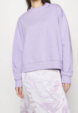 Osoba ubrana w bluzę w pastelowym fioletowym kolorze z prążkowanymi mankietami i dołem, zestawioną z białą spódnicą ozdobioną jasnofioletowym, marmurkowym wzorem.