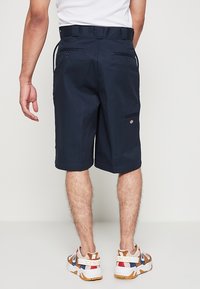Homme portant un short bleu marine mi-genou avec des poches arrière et des baskets multicolores, debout devant un fond clair uni.
