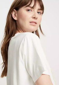 Biała bluzka z krótkim rękawem o swobodnym kroju. Miękka faktura, subtelny połysk i okrągły dekolt. Włosy opadają za ramiona, odkrywając część pleców.