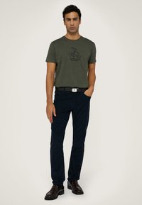 Olijfgroen t-shirt met korte mouwen en een grafische print, gecombineerd met donkerblauwe broek en een zwarte riem met een zilveren gesp. Bruine schoenen.