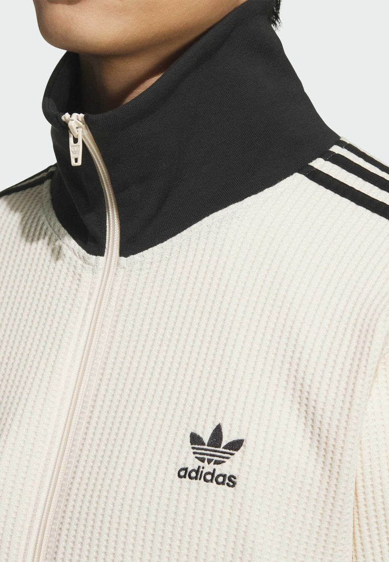 Hvid tekstureret jakke med sort høj krave, med lynlås og tre sorte striber langs skulderen. Adidas-logo på brystet.