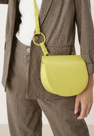 Borsa a tracolla in pelle verde lime di forma rotonda, con tracolla regolabile e dettagli di cucitura in vista, indossata sopra un abbigliamento marrone texturizzato.