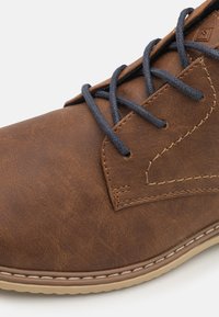 Chaussure en cuir synthétique marron avec une texture lisse, lacets sombres contrastants et accents cousus le long de l'orteil et du côté. Design à bout rond.