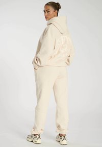Felpa beige con vestibilità oversize, tasche a marsupio e logo stampato sul retro. Abbinata a pantaloni della tuta tapered coordinati e sneaker.