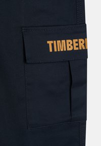 Námornícky modrá látková cargo kapsa s horným klapkovým uzáverom, na ktorom je vodorovne vytlačený výrazný žltý text "TIMBERLAND". Šité okraje a hladká textúra.