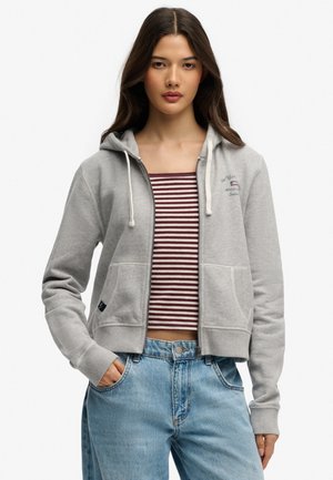Ung kvinde iført en lysegrå hættetrøje med lynlås over en vinrød og hvidstribet crop top og lyseblå jeans, stående med den ene hånd i lommen.