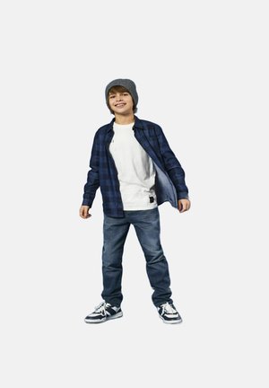 Garçon portant une chemise à carreaux marine par-dessus un T-shirt blanc, un jean bleu et des baskets noir et blanc, avec un bonnet gris et des cheveux en désordre.