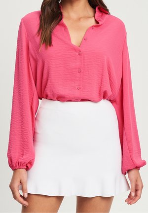 Kvinde iført lyserød langærmet skjorte med knapper, stoppet ned i en hvid mininederdel med svagt flæsekant, stående med afslappede arme.