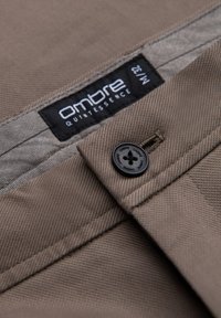 Bruine stoffen broek met diagonale weving, een zwarte knoop en een label met de tekst "ombre QUINTESSENCE N:1/32."