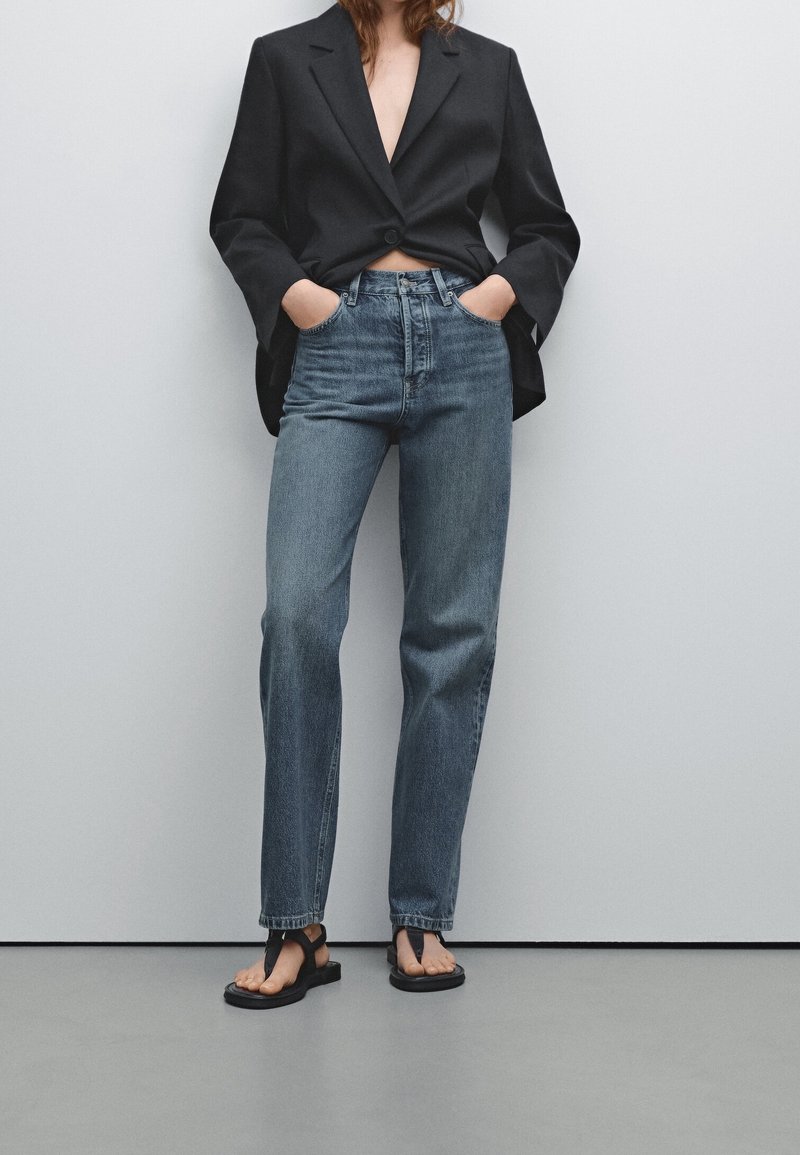 Blazer noir ajusté, jean bleu taille haute et sandales plates noires. Les jeans ont une coupe droite et un léger délavage.