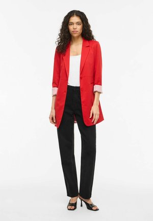 Femme aux cheveux bouclés portant un blazer rouge, un haut blanc, un pantalon noir et des talons ouverts noirs, debout devant un fond blanc uni.