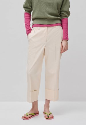 Persona che indossa pantaloni beige a gamba larga e corta, top verdi e rosa a strati e infradito rosa con cinturini verdi, in piedi davanti a uno sfondo grigio.