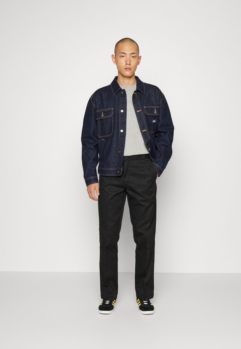 Dark denim jacket with a relaxed fit, featuring gold stitching, two chest pockets, and a gray t-shirt underneath. Worn with black trousers and sneakers.