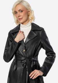 Černý kožený trenčkot s páskem v pase, velkým límcem a knoflíkovým zapínáním. Má hladký povrch a strukturovaný design.