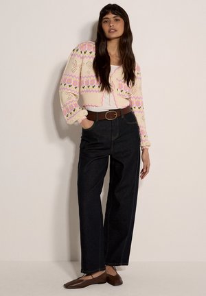 Gestrickte Strickjacke in Creme mit mehrfarbigen Blumenmustern, kombiniert mit hoch taillierten dunklen Jeanshosen und braunen flachen Schuhen.