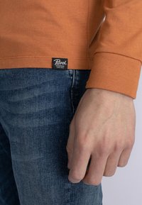 Långärmad orange skjorta i bomull med en strukturerad yta, parat med blå denimjeans. Svart märkesetikett synlig på skjortans fåll.