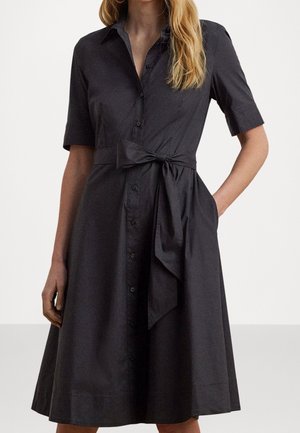 Femme portant une robe noire boutonnée jusqu'aux genoux, à manches courtes et ceinturée à la taille, debout avec une main dans la poche.