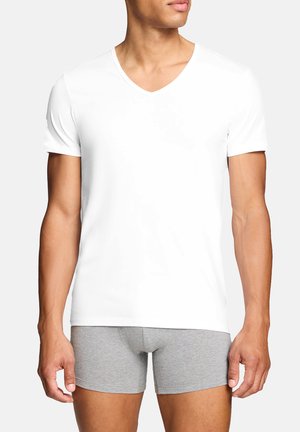 Mann trägt ein schlichtes weißes V-Neck-T-Shirt und graue Boxershorts, steht vor einem einfarbigen hellen Hintergrund.