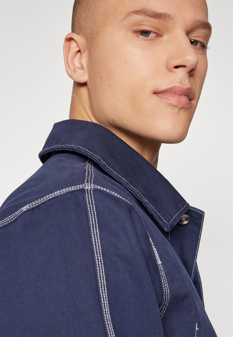 Camisa de mezclilla azul con costuras blancas, un cuello puntiagudo y cierre de botones. La tela parece resistente con un acabado ligeramente texturizado.