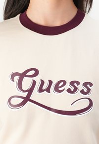 Beige t-skjorte med en burgunder ribbet hals, med en stor, stilisert hvit og burgunder "Guess" tekstlogo over forsiden.